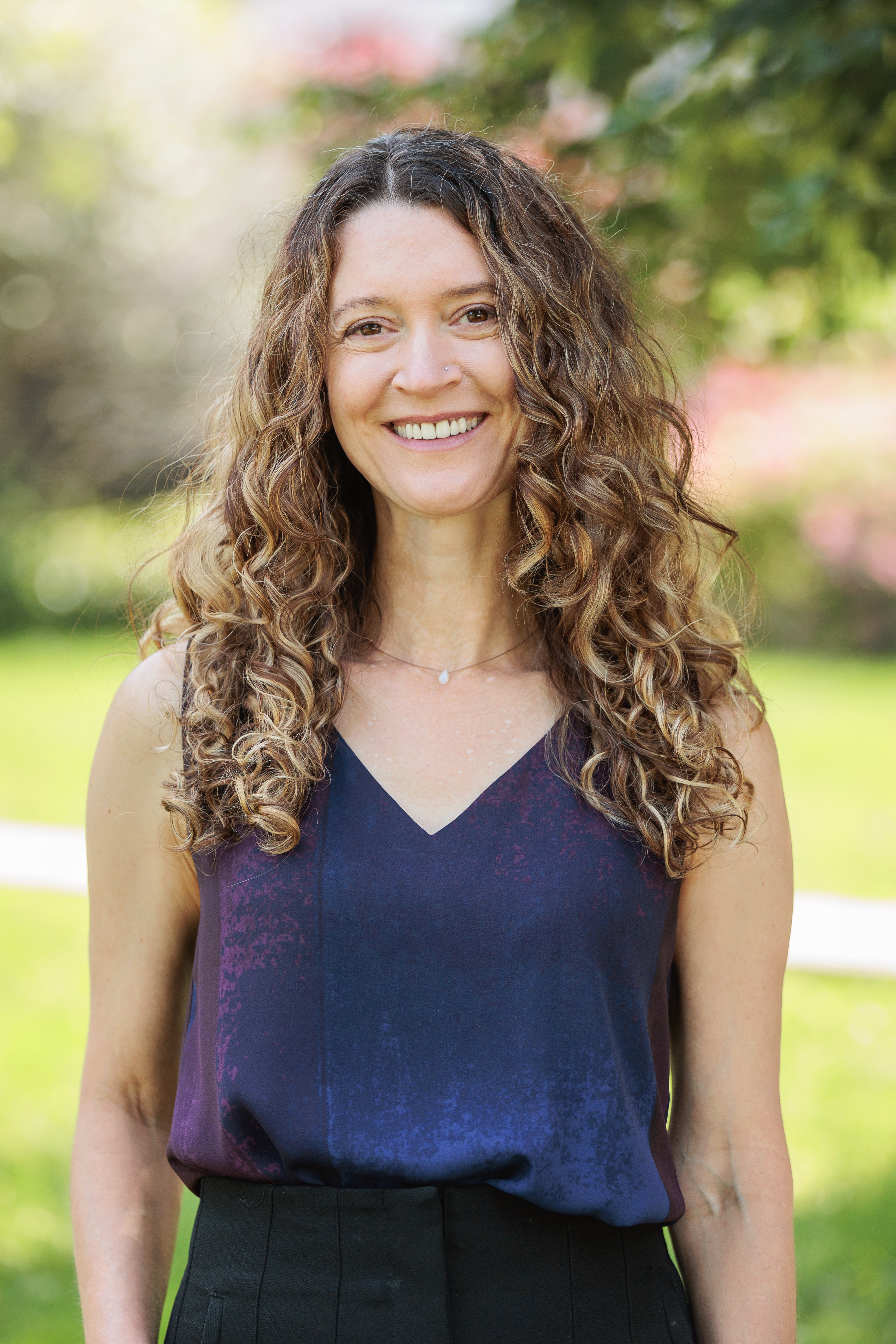 Miché Jo-Keeling, MSW RSW. She is smiling and facing the camera. Her long wavy brown hair touches below her shoulders and she wears a purple v-neck tank blouse and black pants. The background is a soft green.